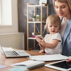 Kobieta z dzieckiem na kolanach pracuje przy biurku. Czy na macierzyńskim można pracować? Tak, nawet z maluchem u boku.