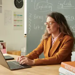 Nauczycielka w okularach pracuje przy laptopie, przygotowując się do awansu zawodowego. Na tablicy napisy po angielsku.