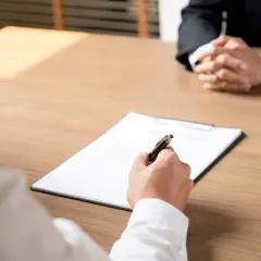 Rozmowa o zwolnieniach grupowych i odprawie. Dłoń pisze na dokumencie, druga osoba siedzi z rękami złożonymi.