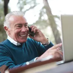 Uśmiechnięty starszy pan rozmawia przez telefon, wskazując na ekran laptopa. Cieszy się okresem ochronnym przed emeryturą, planując przyszłość.