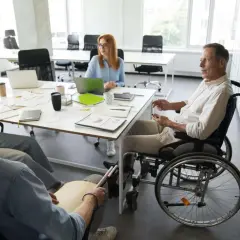 Zespół omawia projekty. Mężczyzna na wózku inwalidzkim i osoba z tabliczką Braille'a, pokazując, że **2 grupa inwalidzka przywileje w pracy** są uwzględniane.