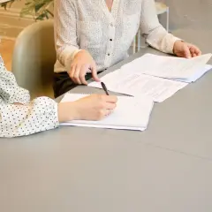 Dwie kobiety omawiają umowę o świadczenie usług. Jedna pisze notatki, druga przegląda dokumenty.