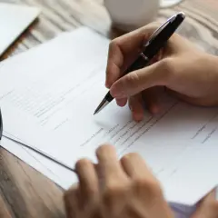 Ręka pisze długopisem na dokumencie, który może być umową o rozwiązanie umowy za porozumieniem stron. Obok leżą okulary.