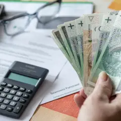 Ręka trzyma polskie banknoty, obok kalkulator, okulary i telefon. Czy to odpowiedź na pytanie, ile wynosi kuroniówka?