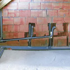Instalacja odpowietrzenia kanału sanitarnego w domu parterowym. Rury PVC biegną wzdłuż czerwonej ściany z pustaków.