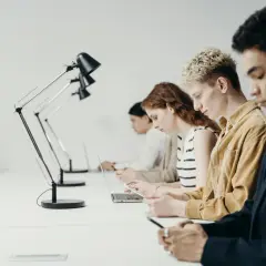 Młodzi ludzie pracują przy biurkach. Zastanawiają się, czy staż jest płatny, skupieni na swoich laptopach i telefonach.