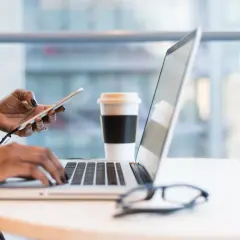 Kobieta pracuje na laptopie, trzymając telefon. Obok stoi kubek kawy. To idealny moment na analizę wniosków o pomoc de minimis.