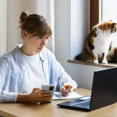 Kobieta w okularach pracuje zdalnie okazjonalnie, notując coś w zeszycie, z telefonem w dłoni. Kot obserwuje świat za oknem.