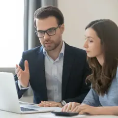 Mężczyzna w okularach tłumaczy kobiecie, jak obliczyć koszty uzyskania przychodu za niepełny miesiąc, patrząc na laptopa.