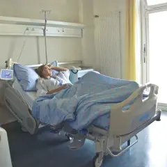 Pacjentka w szpitalnym łóżku, zastanawia się, zwolnienie lekarskie ile płatne. Jasne okno i zasłony.
