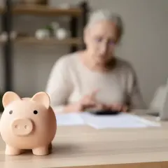 Świnka skarbonka na pierwszym planie, za nią starsza kobieta liczy coś na kalkulatorze. Koniec nadziei na emerytury stażowe?