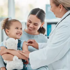 Uśmiechnięta dziewczynka z misiem dostaje szczepienie. Mama wspiera ją, a lekarka delikatnie podaje zastrzyk. To ważny element opieki nad dzieckiem.