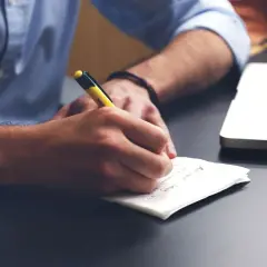Mężczyzna w niebieskiej koszuli pisze coś długopisem na kartce. Obok leży laptop. To może być korekta świadectwa pracy.