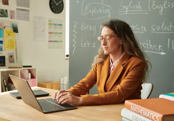 Nauczycielka w okularach pracuje przy laptopie, przygotowując się do awansu zawodowego. Na tablicy napisy po angielsku.