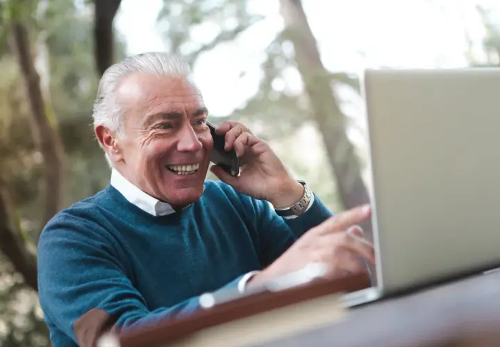 Uśmiechnięty starszy pan rozmawia przez telefon, wskazując na ekran laptopa. Cieszy się okresem ochronnym przed emeryturą, planując przyszłość.