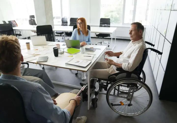 Zespół omawia projekty. Mężczyzna na wózku inwalidzkim i osoba z tabliczką Braille'a, pokazując, że **2 grupa inwalidzka przywileje w pracy** są uwzględniane.