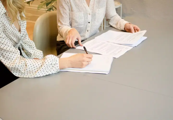 Dwie kobiety omawiają umowę o świadczenie usług. Jedna pisze notatki, druga przegląda dokumenty.