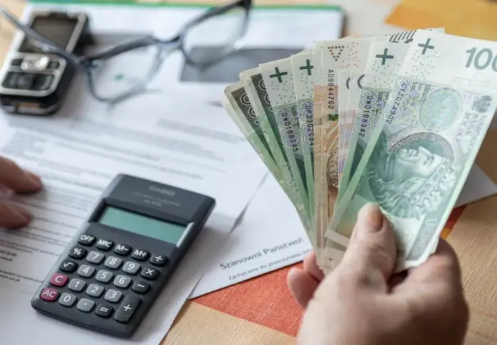 Ręka trzyma polskie banknoty, obok kalkulator, okulary i telefon. Czy to odpowiedź na pytanie, ile wynosi kuroniówka?