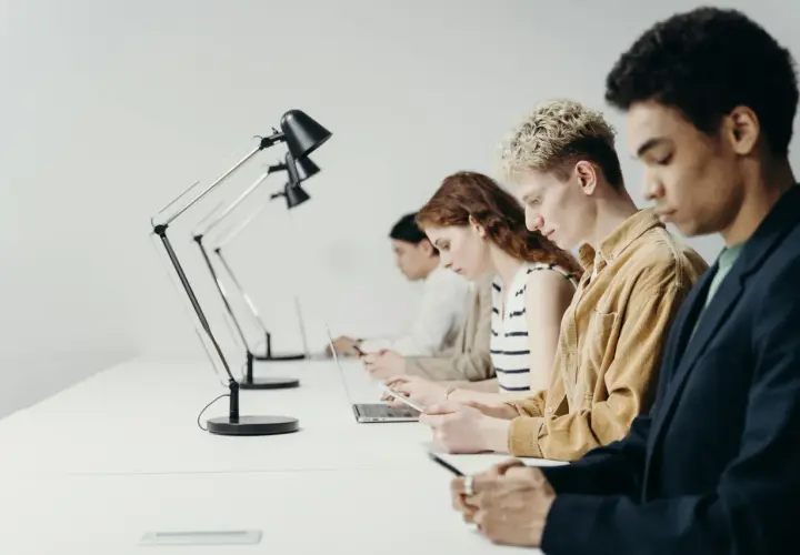 Młodzi ludzie pracują przy biurkach. Zastanawiają się, czy staż jest płatny, skupieni na swoich laptopach i telefonach.