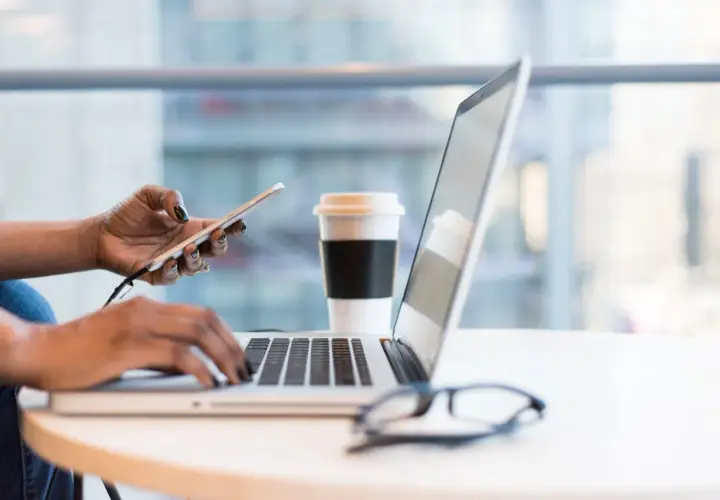 Kobieta pracuje na laptopie, trzymając telefon. Obok stoi kubek kawy. To idealny moment na analizę wniosków o pomoc de minimis.