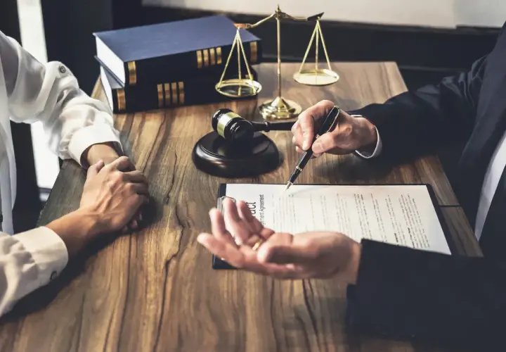 Prawnik omawia z klientem dokumenty, wskazując piórem na umowę. W tle waga sprawiedliwości i księgi. To ważny moment, by złożyć list intencyjny praca.