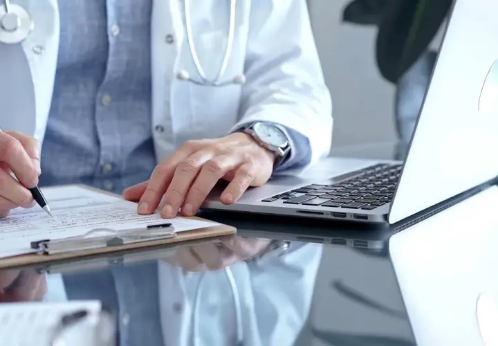 Lekarz w białym kitlu pisze na dokumentach, obok laptop. Zastanawia się, ile można być na zwolnieniu lekarskim od psychiatry.