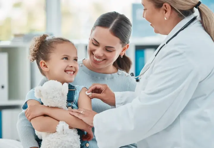 Uśmiechnięta dziewczynka z misiem dostaje szczepienie. Mama wspiera ją, a lekarka delikatnie podaje zastrzyk. To ważny element opieki nad dzieckiem.