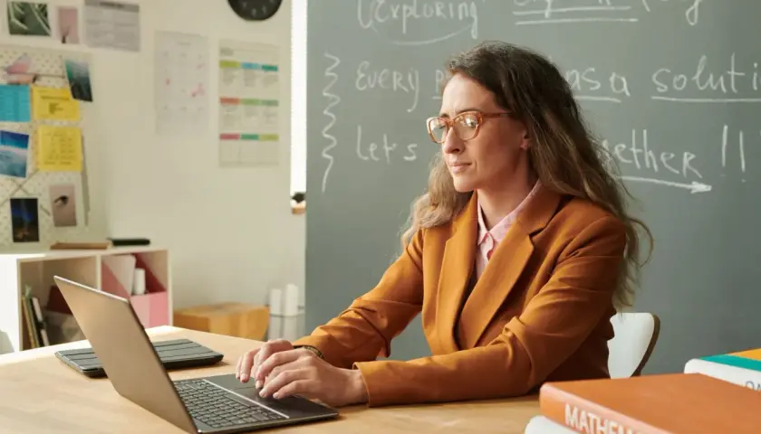 Nauczycielka w okularach pracuje przy laptopie, przygotowując się do awansu zawodowego. Na tablicy napisy po angielsku.