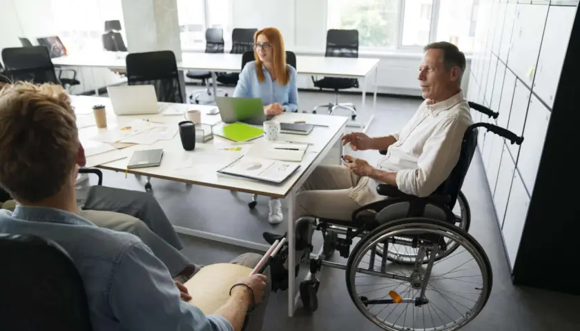 Zespół omawia projekty. Mężczyzna na wózku inwalidzkim i osoba z tabliczką Braille'a, pokazując, że **2 grupa inwalidzka przywileje w pracy** są uwzględniane.