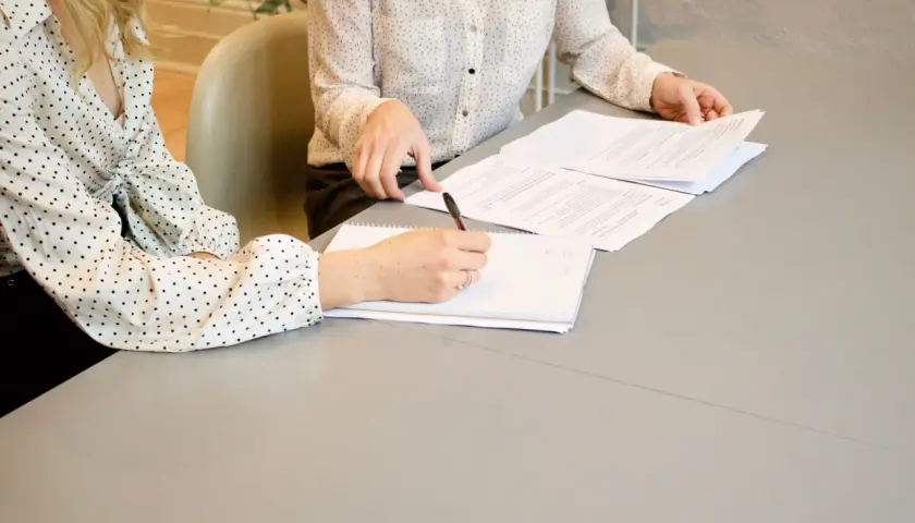 Dwie kobiety omawiają umowę o świadczenie usług. Jedna pisze notatki, druga przegląda dokumenty.