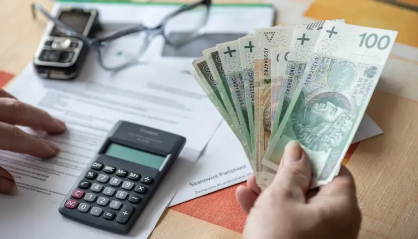 Ręka trzyma polskie banknoty, obok kalkulator, okulary i telefon. Czy to odpowiedź na pytanie, ile wynosi kuroniówka?