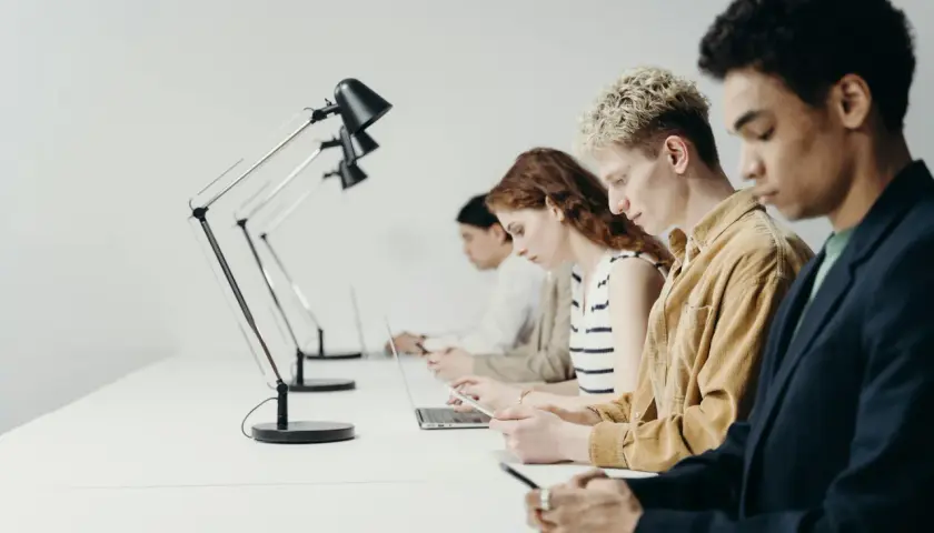 Młodzi ludzie pracują przy biurkach. Zastanawiają się, czy staż jest płatny, skupieni na swoich laptopach i telefonach.