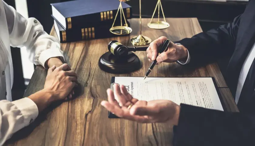Prawnik omawia z klientem dokumenty, wskazując piórem na umowę. W tle waga sprawiedliwości i księgi. To ważny moment, by złożyć list intencyjny praca.
