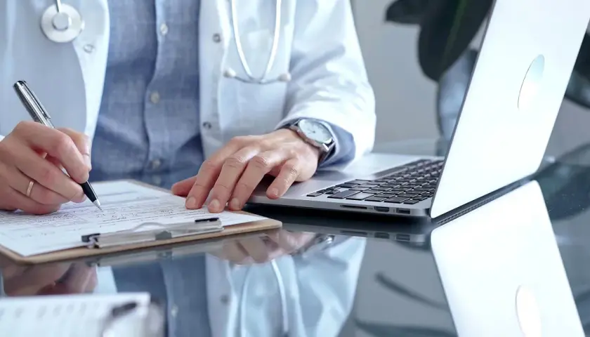 Lekarz w białym kitlu pisze na dokumentach, obok laptop. Zastanawia się, ile można być na zwolnieniu lekarskim od psychiatry.