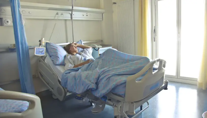 Pacjentka w szpitalnym łóżku, zastanawia się, zwolnienie lekarskie ile płatne. Jasne okno i zasłony.