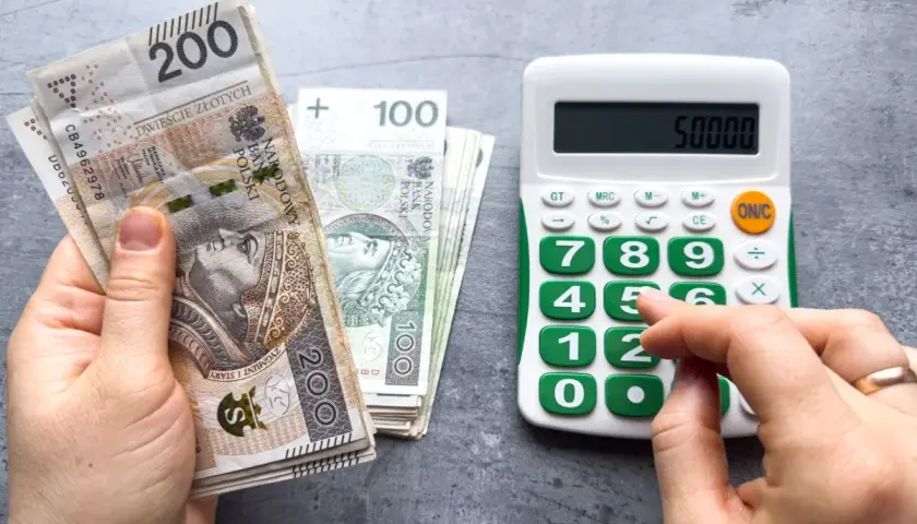 Dłoń trzyma banknoty 200 zł i 100 zł, obok kalkulator z wyświetloną kwotą 50000. Może to być podstawa do obliczenia składek ZUS, np. kod 0590.