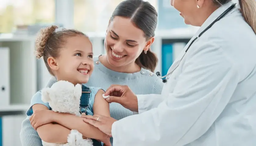 Uśmiechnięta dziewczynka z misiem dostaje szczepienie. Mama wspiera ją, a lekarka delikatnie podaje zastrzyk. To ważny element opieki nad dzieckiem.
