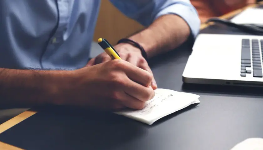 Mężczyzna w niebieskiej koszuli pisze coś długopisem na kartce. Obok leży laptop. To może być korekta świadectwa pracy.