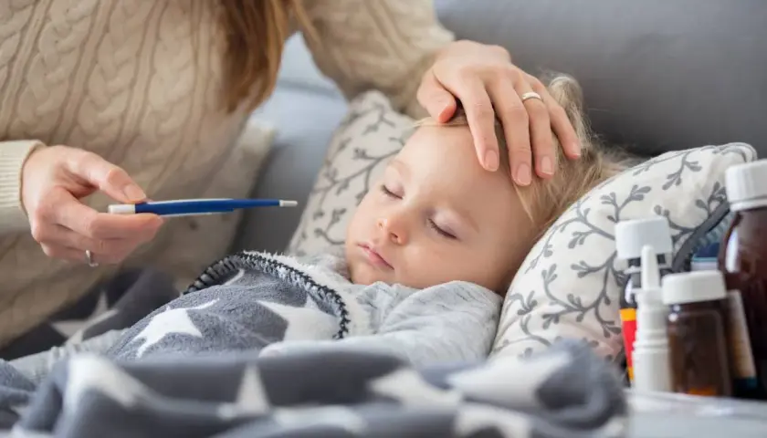 Czuła opieka na dziecko podczas choroby. Mama mierzy gorączkę, głaszcze po czole. Obok leki.