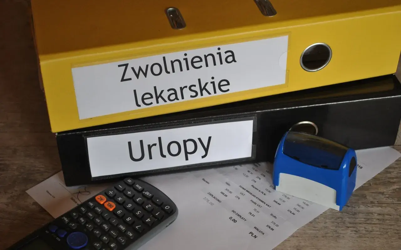 Segregator "Zwolnienia lekarskie" i "Urlopy" obok kalkulatora i pieczątki. Dokumenty mogą zawierać kod choroby a.