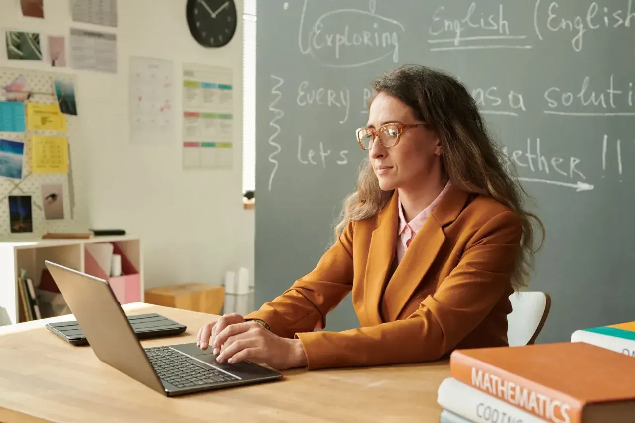 Nauczycielka w okularach pracuje przy laptopie, przygotowując się do awansu zawodowego. Na tablicy napisy po angielsku.
