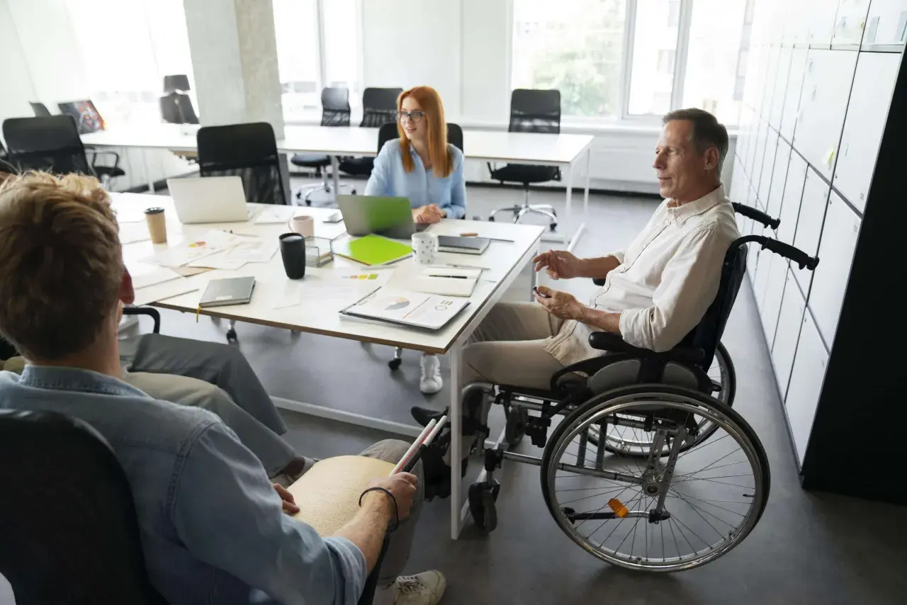 Zespół omawia projekty. Mężczyzna na wózku inwalidzkim i osoba z tabliczką Braille'a, pokazując, że **2 grupa inwalidzka przywileje w pracy** są uwzględniane.