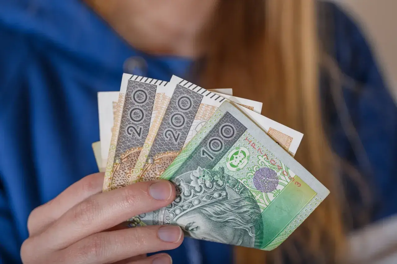 Dłoń trzyma polskie banknoty 100 i 200 zł. To symbol dodatku za wysługę lat w budżetówce.