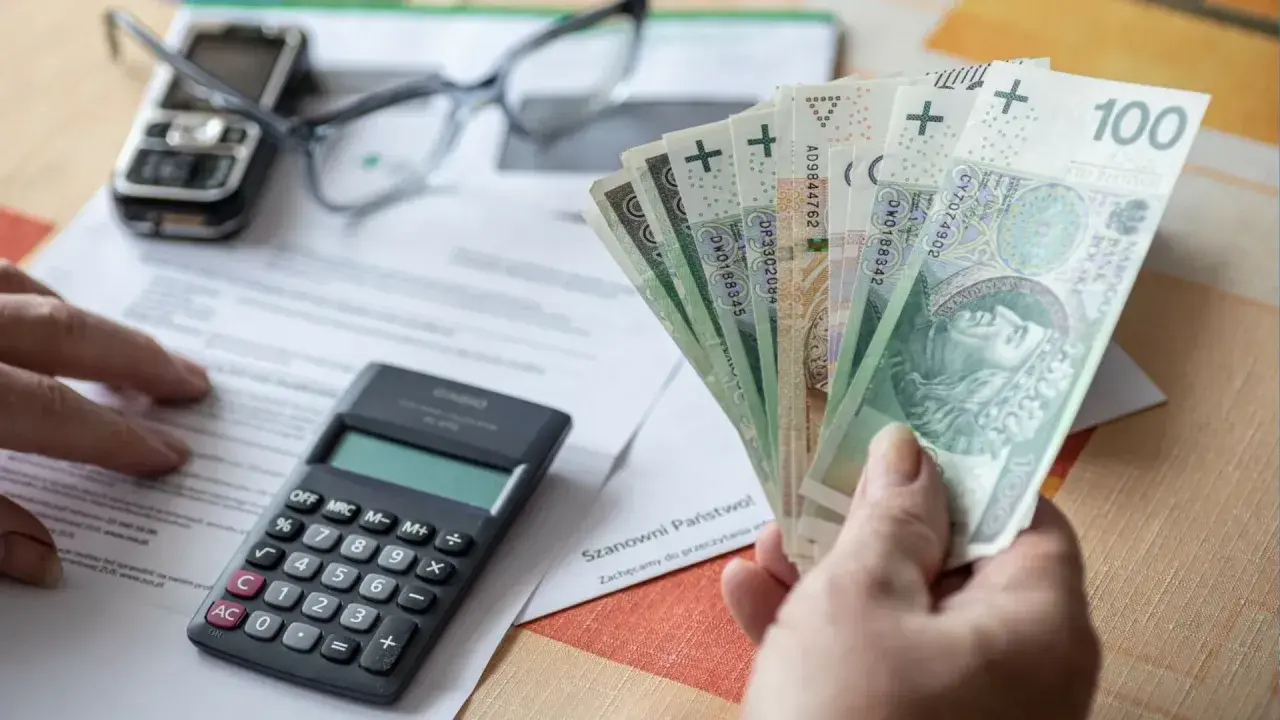 Ręka trzyma polskie banknoty, obok kalkulator, okulary i telefon. Czy to odpowiedź na pytanie, ile wynosi kuroniówka?