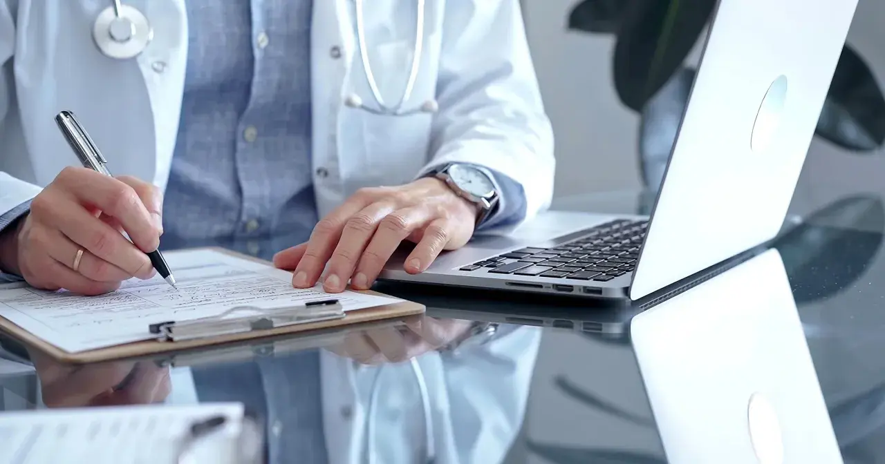 Lekarz w białym kitlu pisze na dokumentach, obok laptop. Zastanawia się, ile można być na zwolnieniu lekarskim od psychiatry.