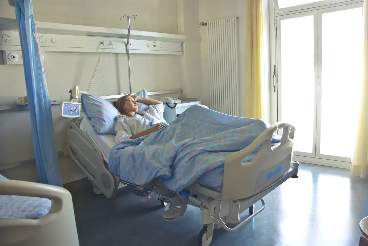 Pacjentka w szpitalnym łóżku, zastanawia się, zwolnienie lekarskie ile płatne. Jasne okno i zasłony.