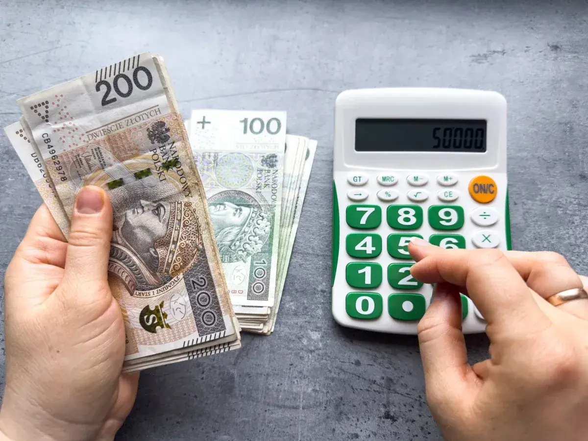 Dłoń trzyma banknoty 200 zł i 100 zł, obok kalkulator z wyświetloną kwotą 50000. Może to być podstawa do obliczenia składek ZUS, np. kod 0590.