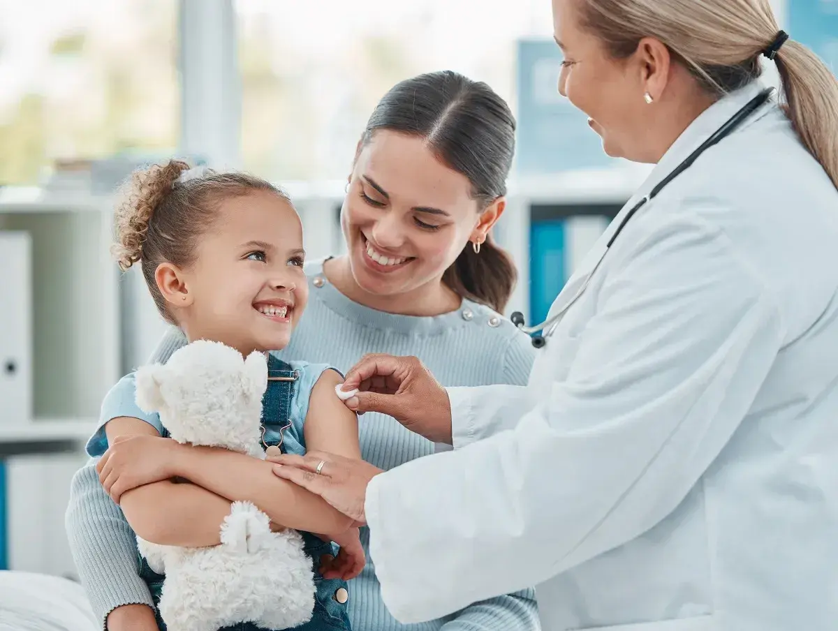 Uśmiechnięta dziewczynka z misiem dostaje szczepienie. Mama wspiera ją, a lekarka delikatnie podaje zastrzyk. To ważny element opieki nad dzieckiem.