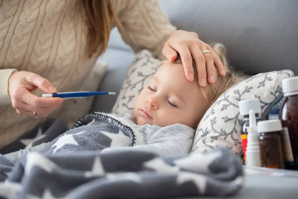 Czuła opieka na dziecko podczas choroby. Mama mierzy gorączkę, głaszcze po czole. Obok leki.