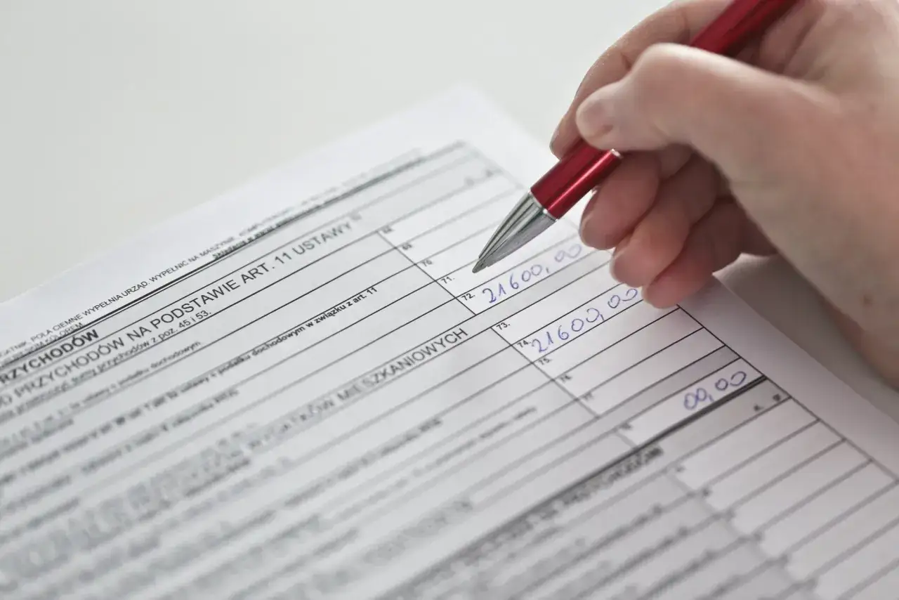 Dłoń z czerwonym długopisem wypełnia rubryki formularza podatkowego, wpisując kwoty. To część rozliczenia przychod&oacute;w na podstawie art. 81 ordynacji podatkowej.